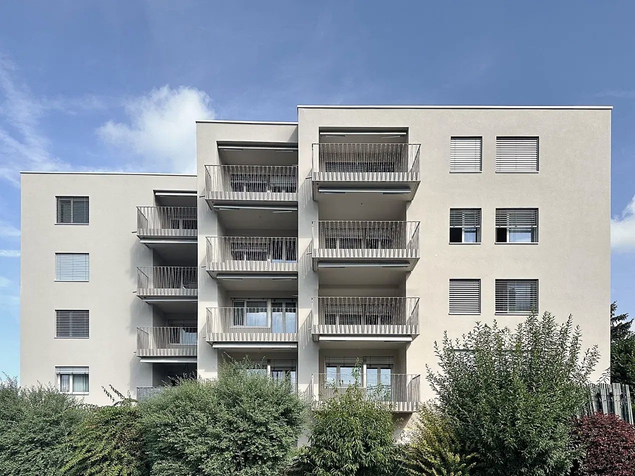 Helle Fassade eines Mehrfamilienhauses mit Balkonen und Lamellenstoren vor blauem Himmel